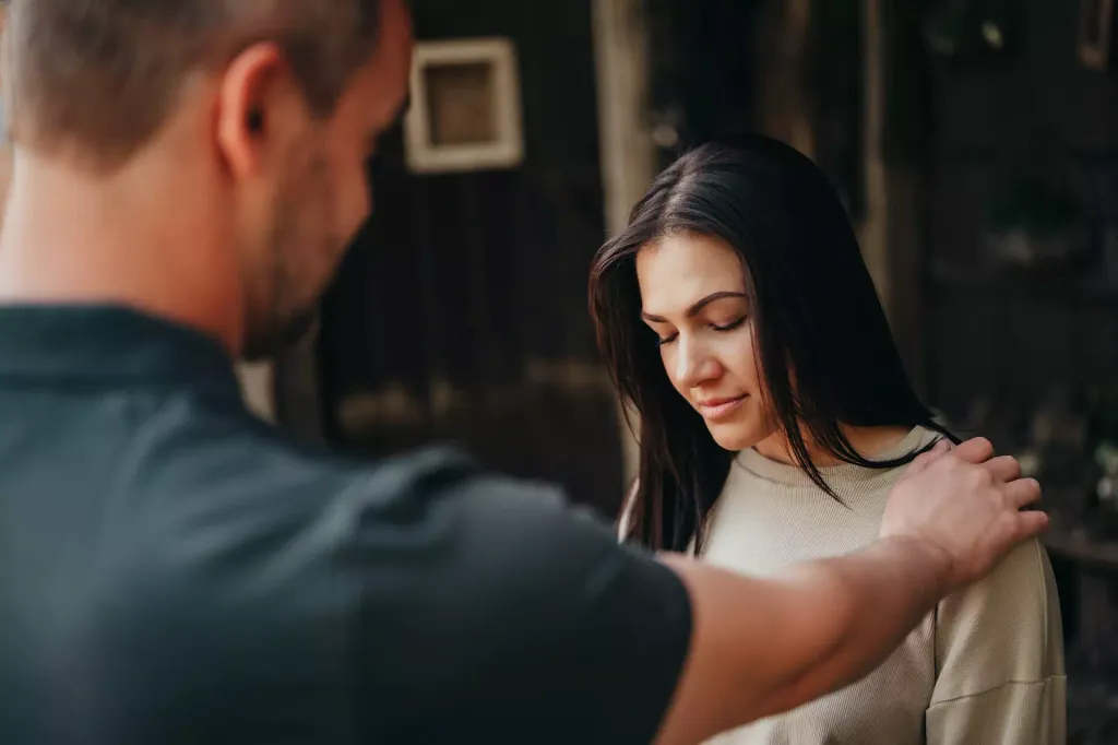 A man places his hand comfortingly on the shoulder of a pensive woman who keeps her eyes downcast