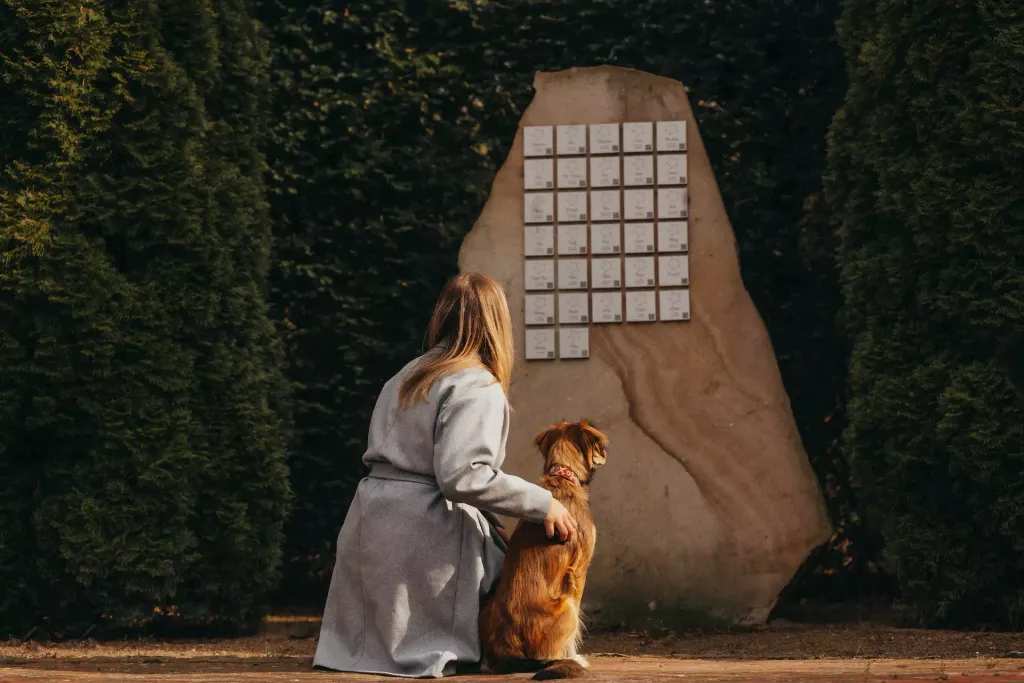 A woman with a dog in front of a memorial stone with name plaques in a peaceful, green setting