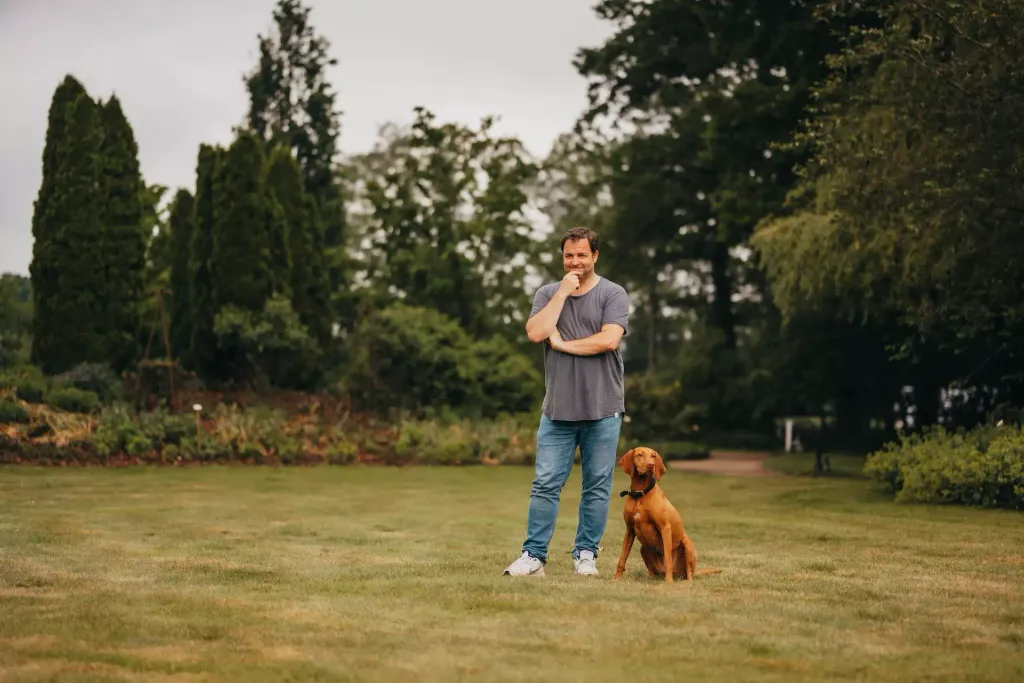 Martin Rütter is standing on a lawn in a well-kept park. A dog is sitting next to him.